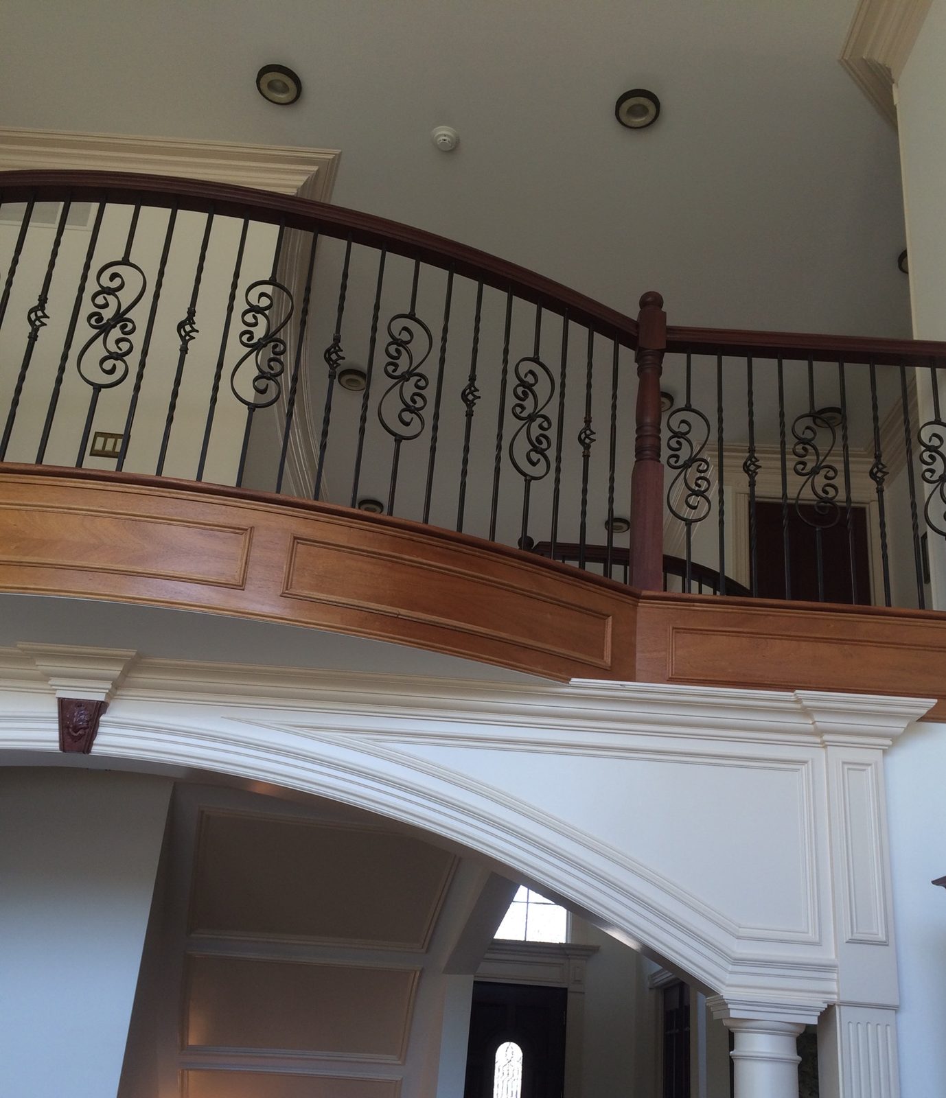 Painted arch with a keystone and a column under a curved, paneled balcony with iron balusters, and a paneled barrel ceiling beyond it.