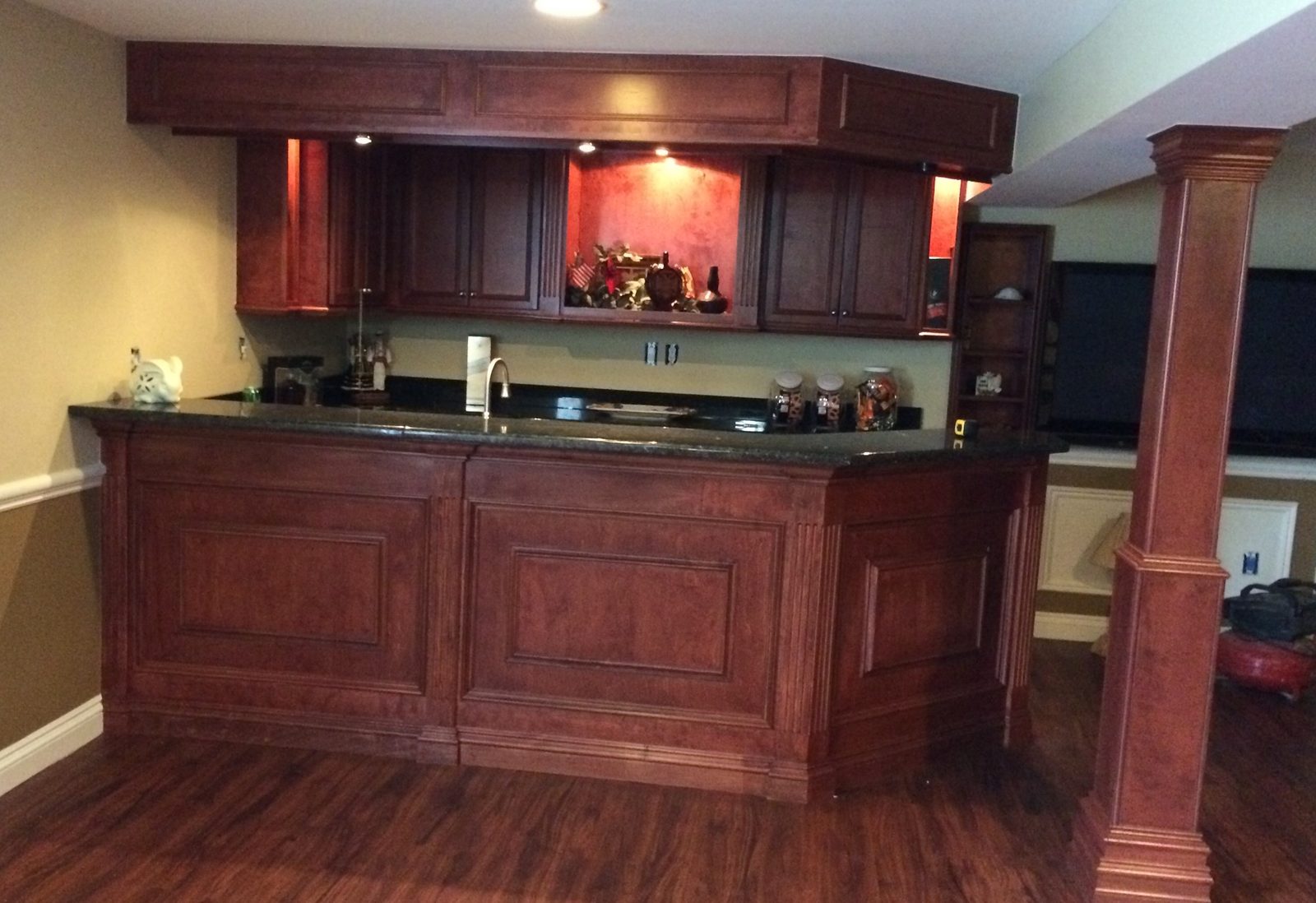 Finished basement bar in stained wood: raised-panel front, fluted pilasters, a paneled soffit with lights over the wall cabinets, and a wrapped column.
