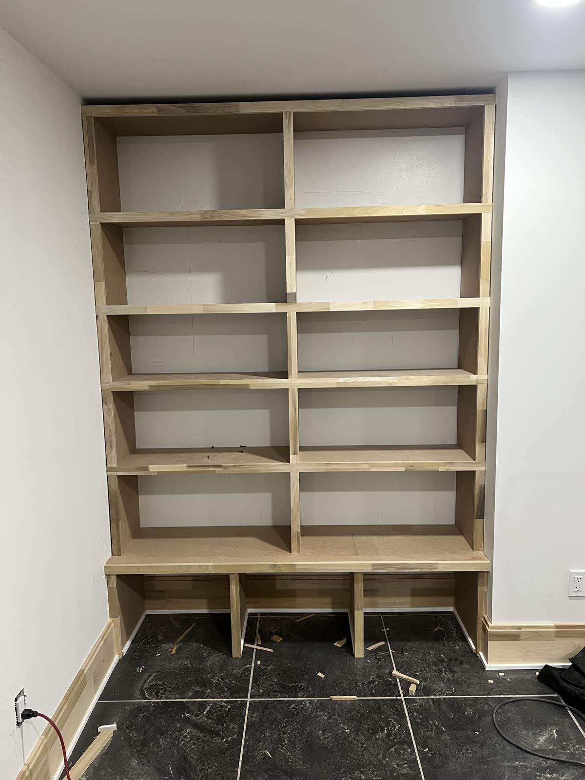Floor-to-ceiling bookcase before paint, raw wood face frames and MDF, two columns of shelves over an open base with dividers, on dark tile.