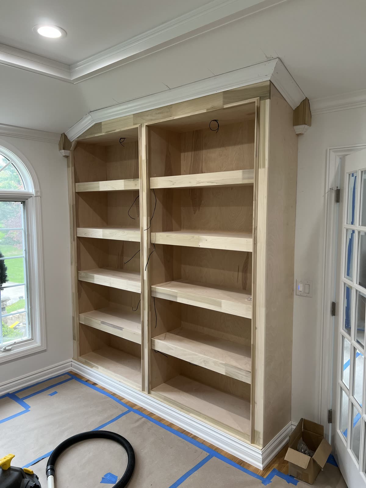 Two-bay bookcase before paint, raw wood, crown molding at a tray ceiling and wires hanging through the back panels.