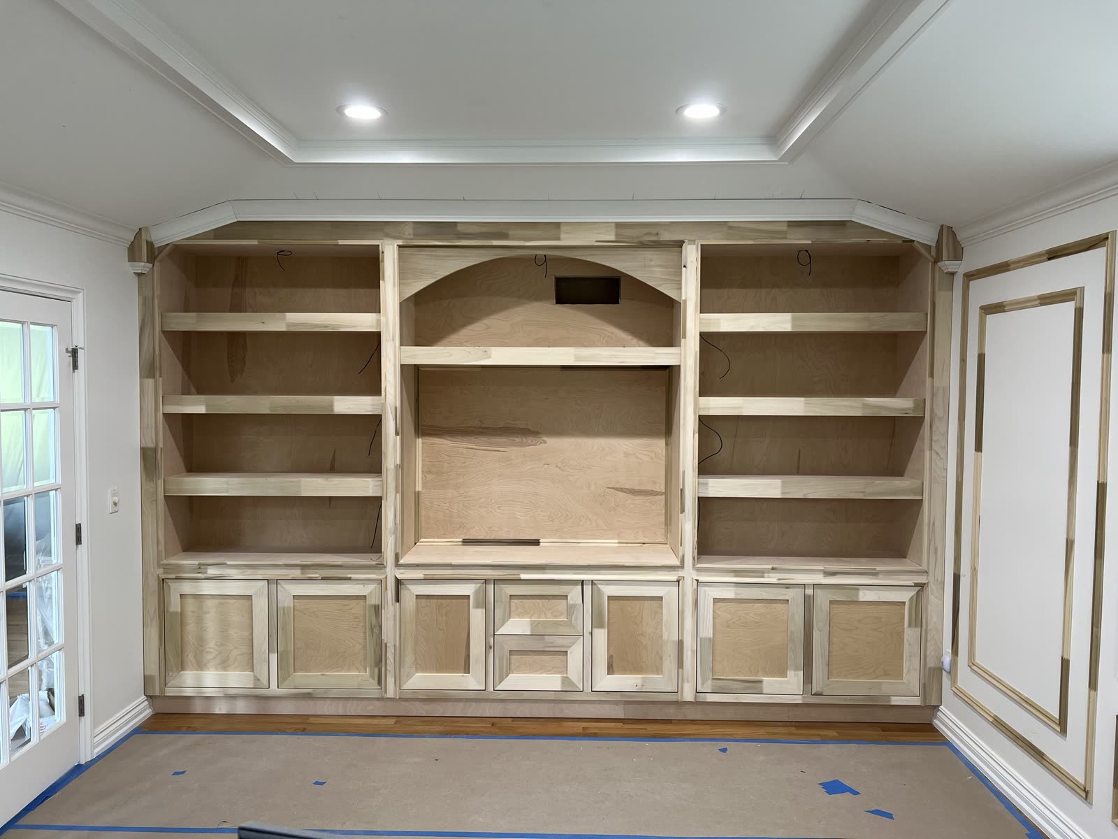 Wall-to-wall built-in before paint, all raw wood: open shelves on both sides of an arched center niche, shaker doors and two drawers along the bottom, crown with angled returns at the ceiling, and nested molding frames on the side wall.