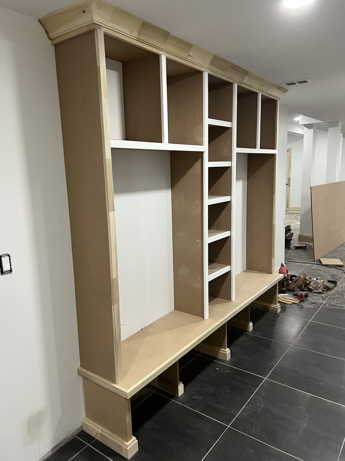 Mudroom bench and locker built-in before paint, seen from an angle: crown at the top, open cubbies, a narrow center column of shelves, and a bench seat on plinth feet, raw wood and MDF on black tile.